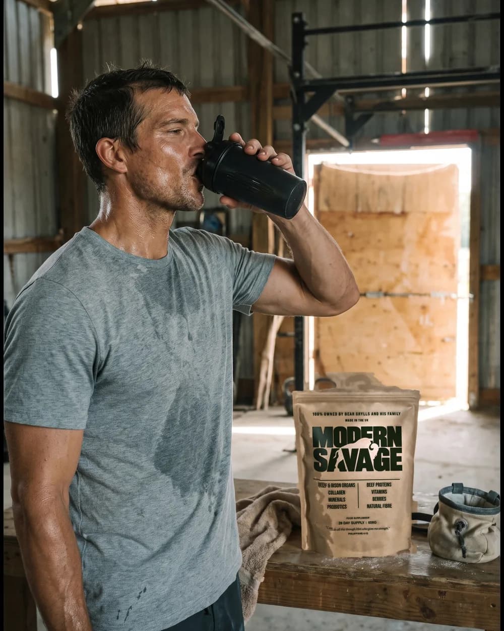 Bear Grylls drinking from a shaker in a barn gym after training, a Modern Savage pouch on the bench behind him