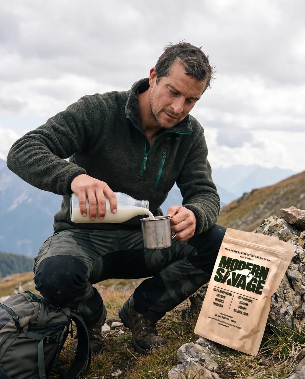 Bear Grylls crouched on a mountain ridge pouring milk into a tin mug, a Modern Savage pouch on the rock beside him