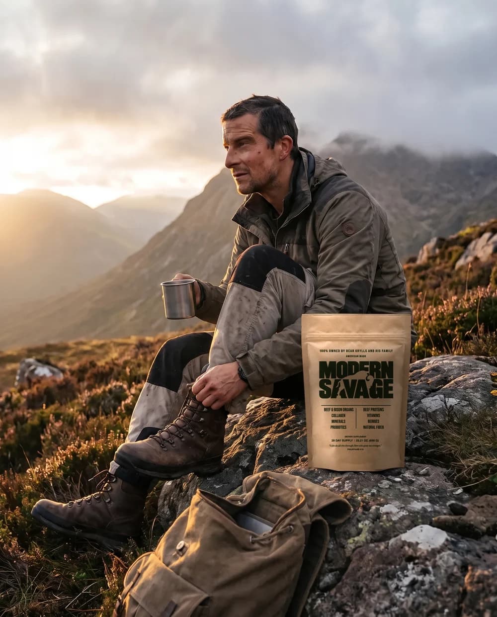 Bear Grylls on a mountain rock at sunrise holding a steel mug, a Modern Savage pouch beside him and a rucksack at his feet