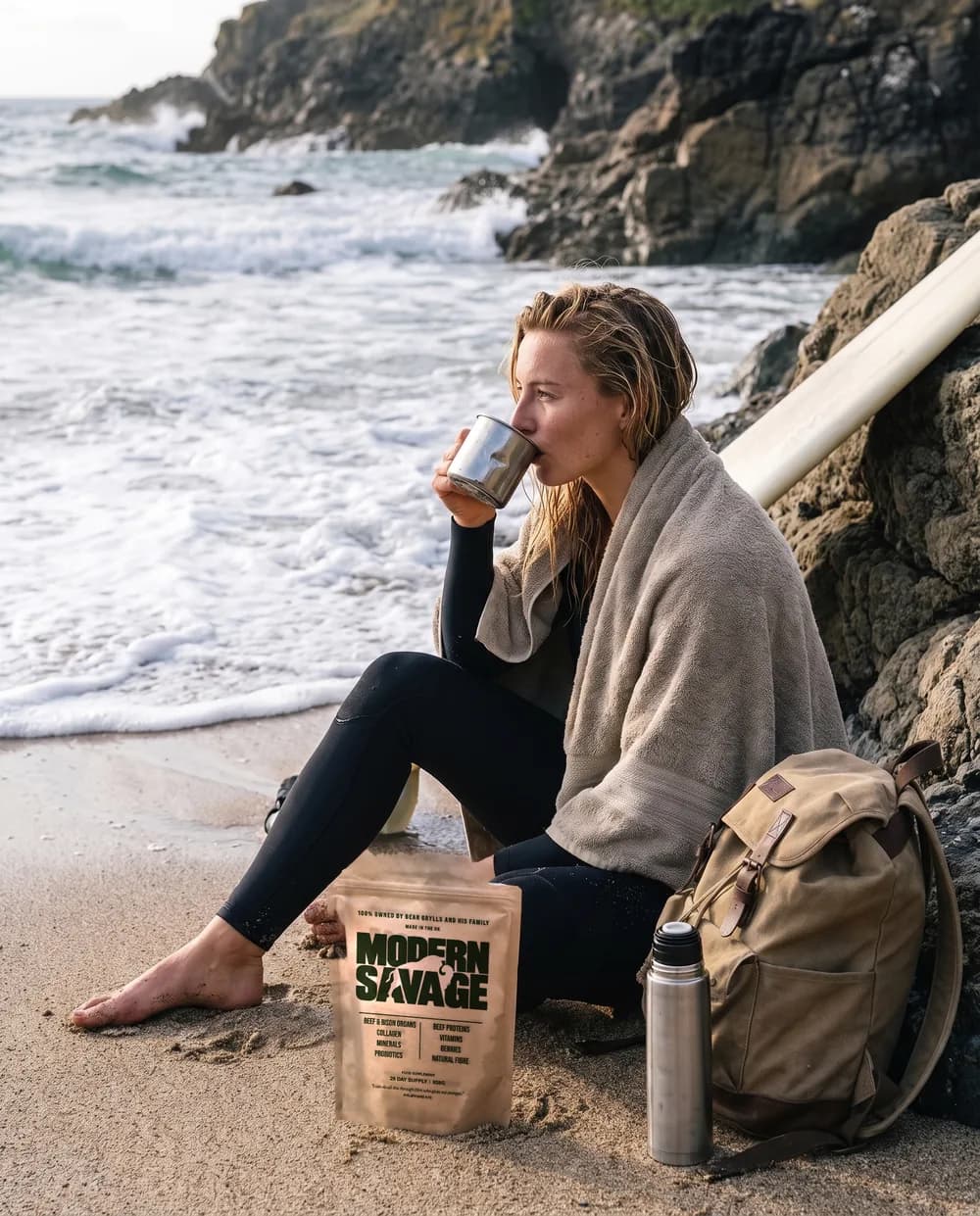 A surfer wrapped in a towel on the sand after a session, drinking from a mug, a Modern Savage pouch by her bag