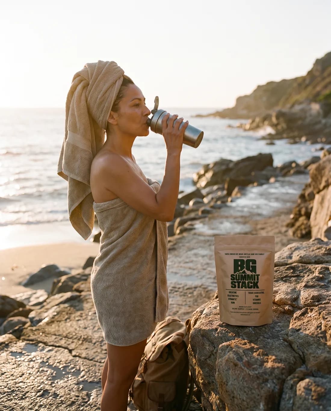 A woman in a towel after a sea swim drinking from a bottle, a BG Summit Stack pouch on a rock